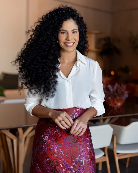 Dra. Sara Dutra em pé, de camisa branca e saia vinho, sorrindo