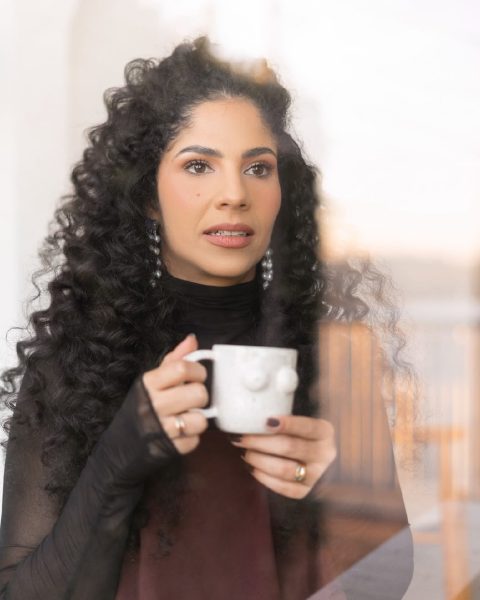 Dra. Sara Dutra segurando uma caneca junto à janela, em tom pensativo