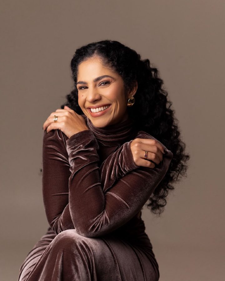 Dra. Sara Dutra sentada, de vestido de veludo marrom, sorrindo com os braços cruzados sobre os ombros
